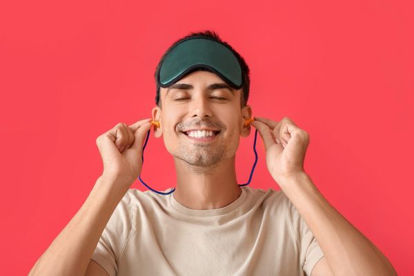 Smiling young man with sleeping mask putting ear plugs on red background